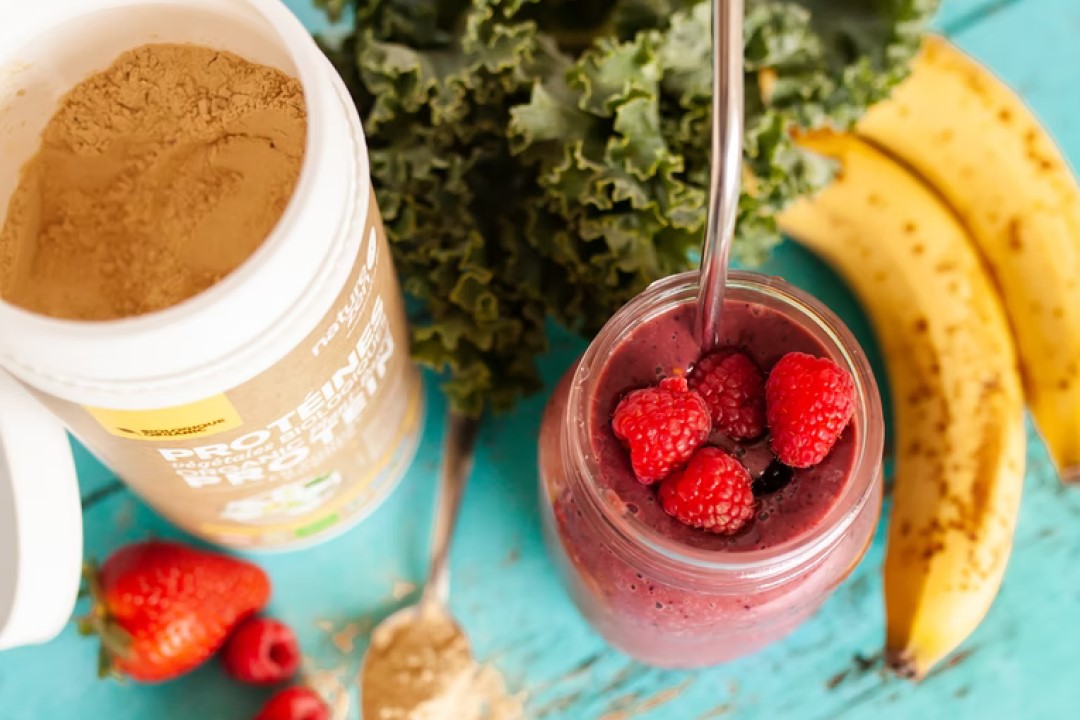 A canister of protein drink powder next to a smoothie glass with berries
